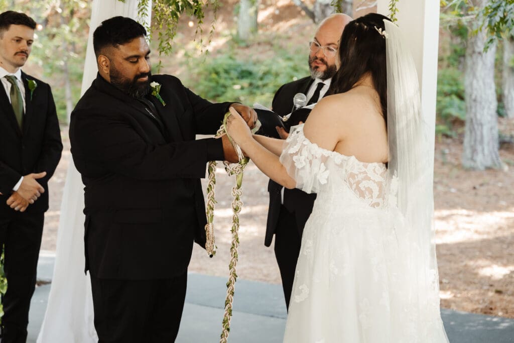 Bride and groom participating in a handfasting ritual during their outdoor fairytale wedding ceremony