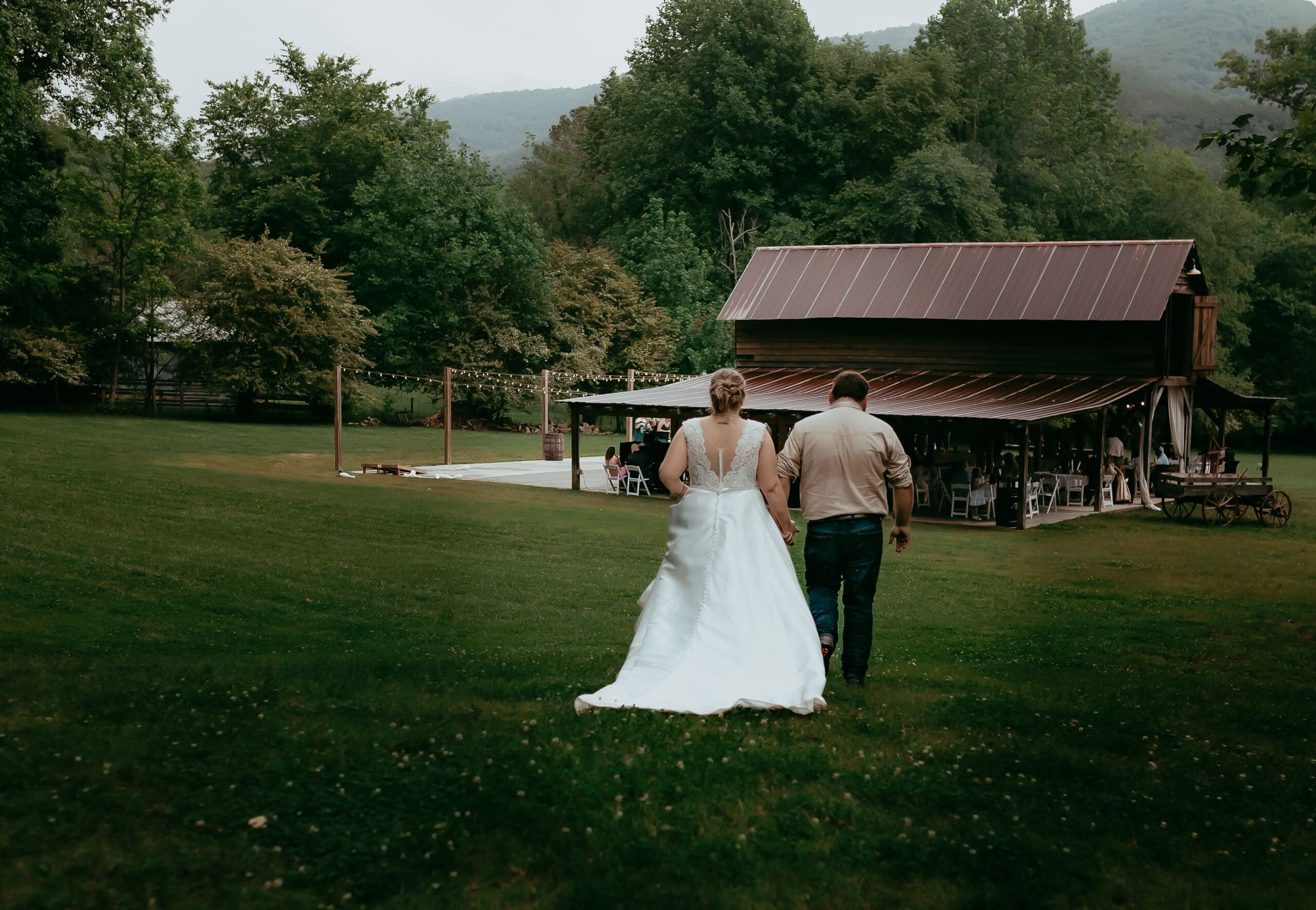 Bride and groom walking hand in hand across a grassy field toward a rustic barn wedding reception in the Georgia mountains, captured in a relaxed documentary style.