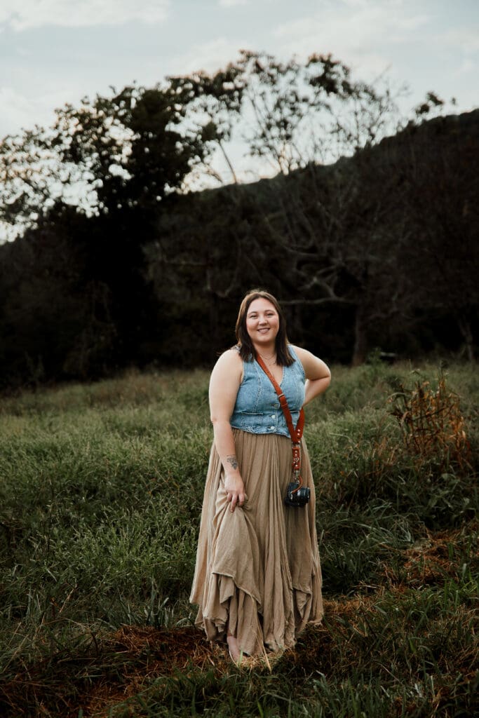 Photographer poses with camera in grassy meadow during an overcast day.