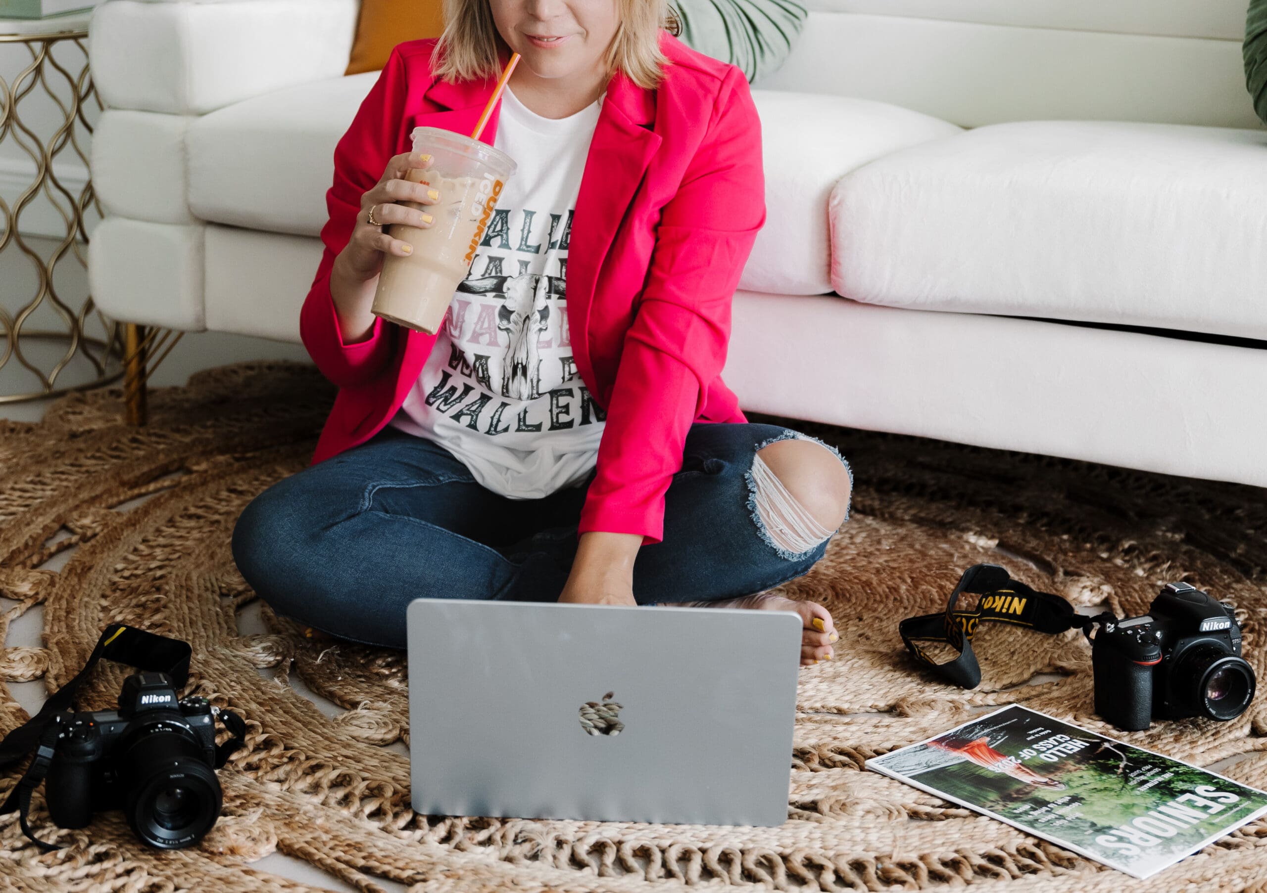 Branding photographer working on a laptop while seated on the floor, holding iced coffee with cameras and creative tools nearby