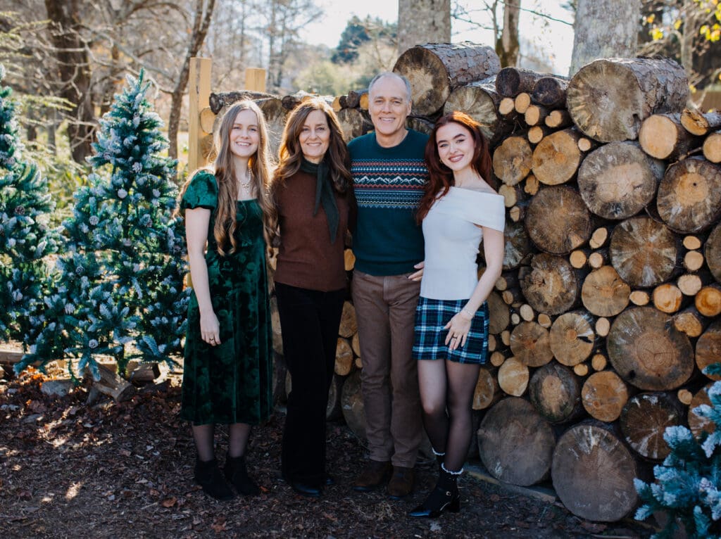 Lifestyle family Christmas session captured by a Georgia family photographer with parents and daughters standing together beside rustic wood stacks and evergreen trees.
