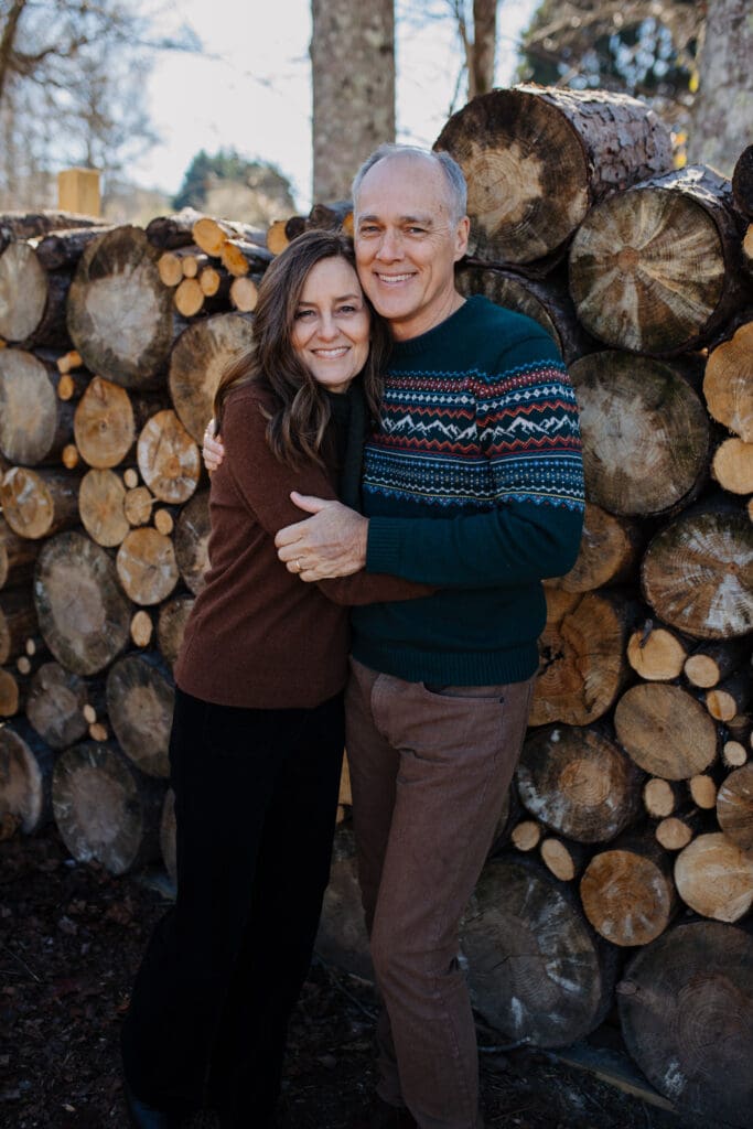 Romantic Christmas couple portrait photographed by a Georgia family photographer with parents hugging in front of stacked firewood in a natural winter setting.