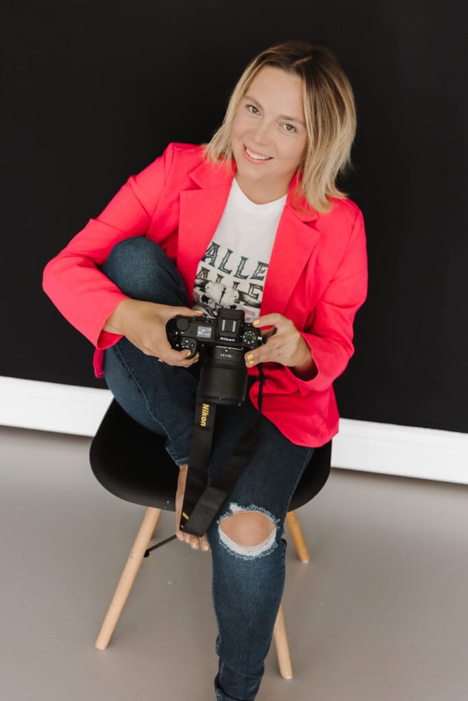 Photographer smiling while seated on a stool, holding a camera against a dark studio backdrop
