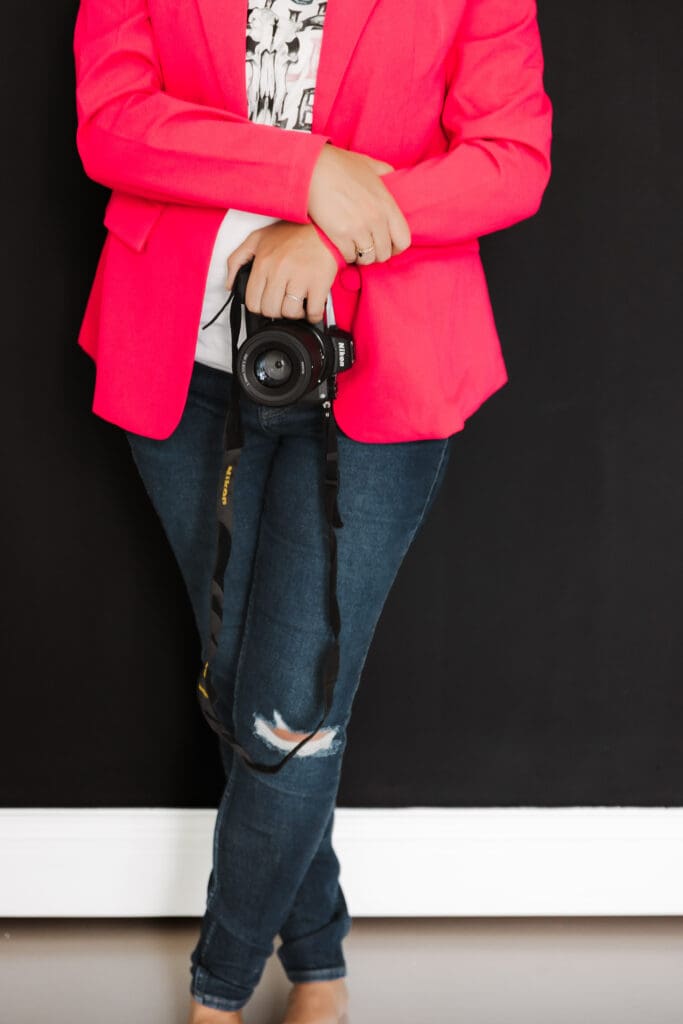 Close-up detail of a branding photographer holding a camera, wearing a bright pink blazer and casual denim