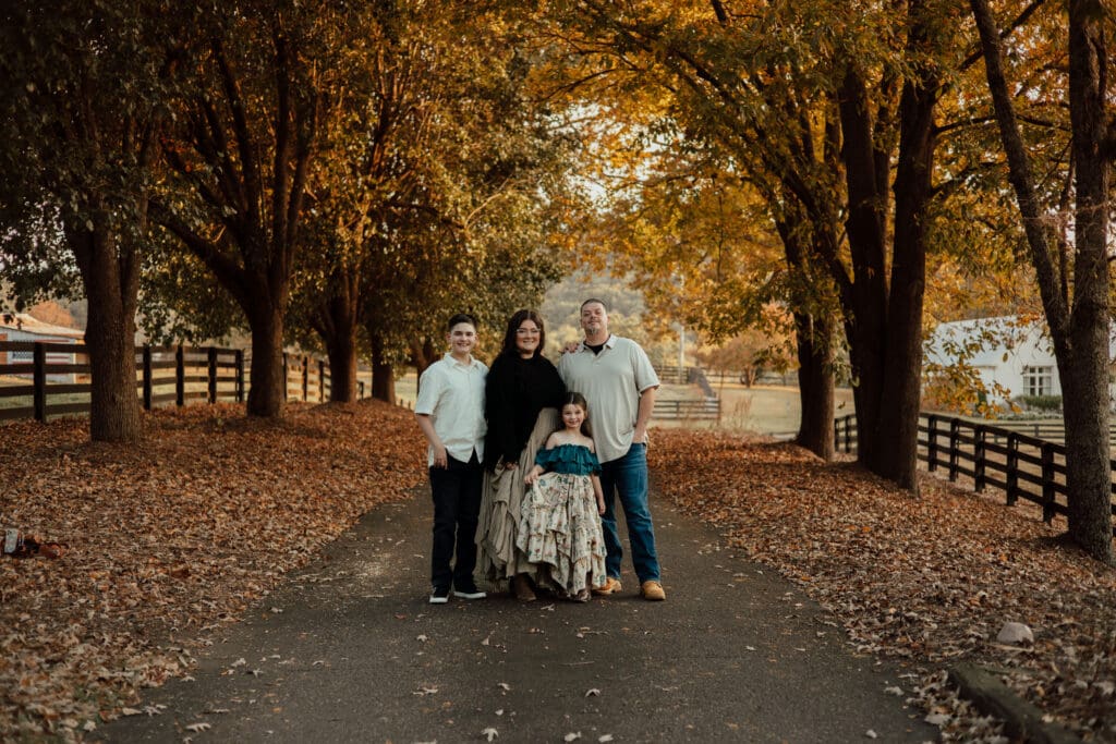Family of four standing together on a tree-lined road during fall in North Georgia, surrounded by golden autumn leaves