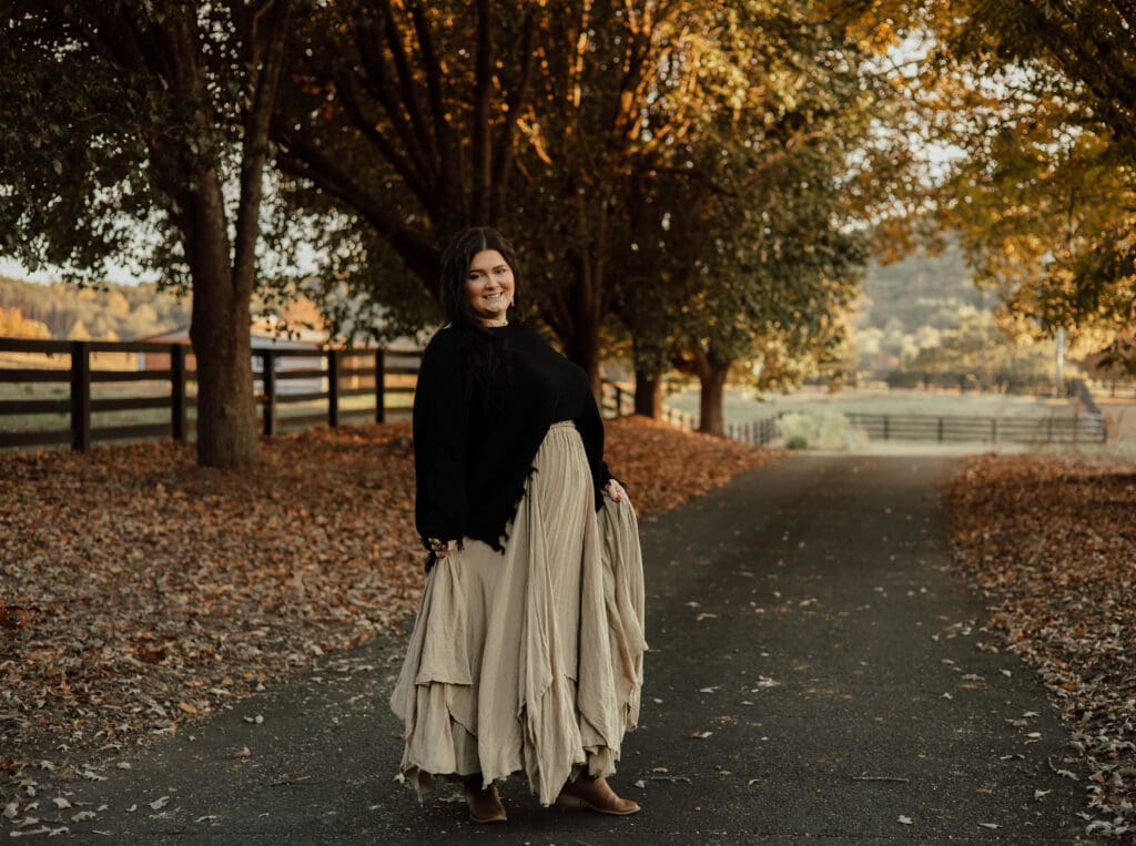 Mother standing on a tree-lined path during a family photo session, surrounded by fall foliage at golden hour.