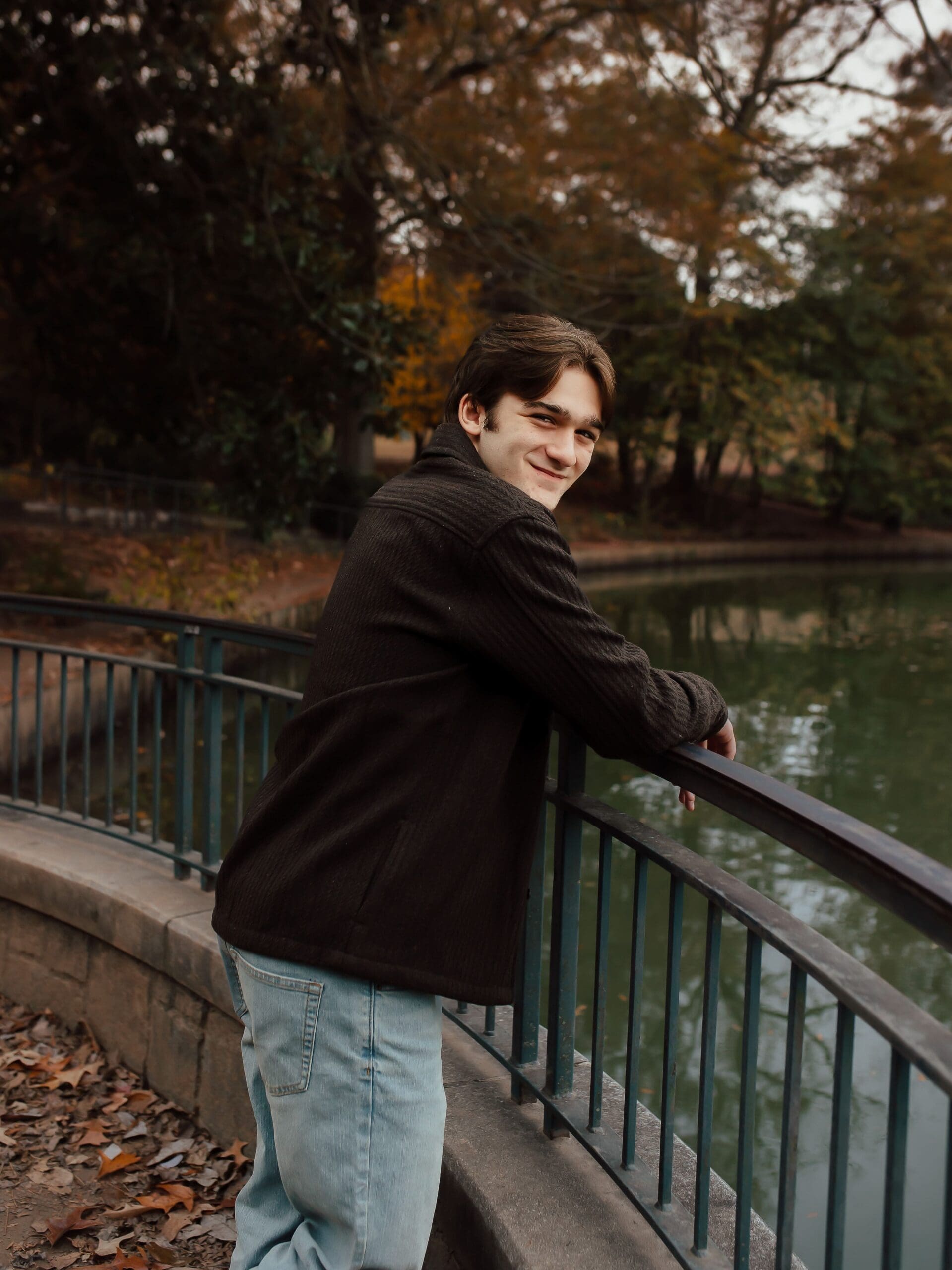 Senior boy leaning against a railing at Piedmont Park with fall trees and water behind him during an Atlanta senior session