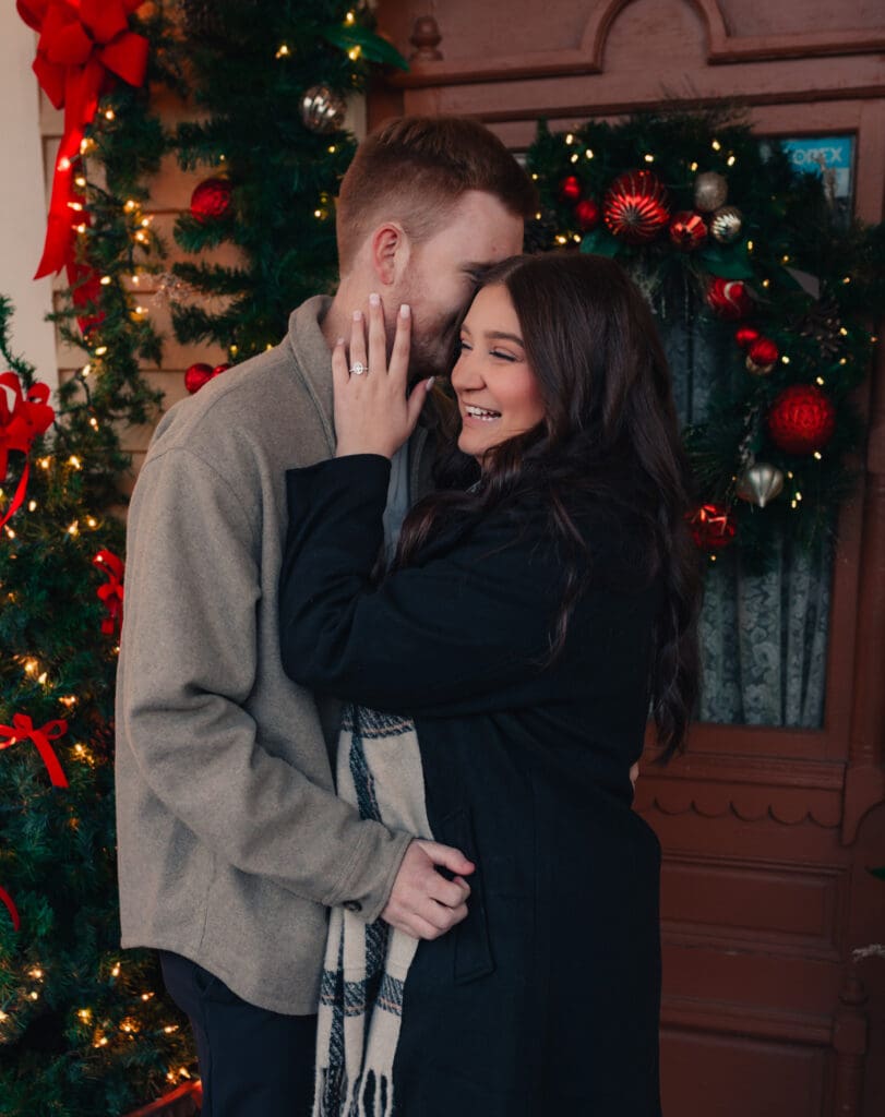 Romantic engagement portrait of a couple laughing together in front of holiday greenery and glowing Christmas decor