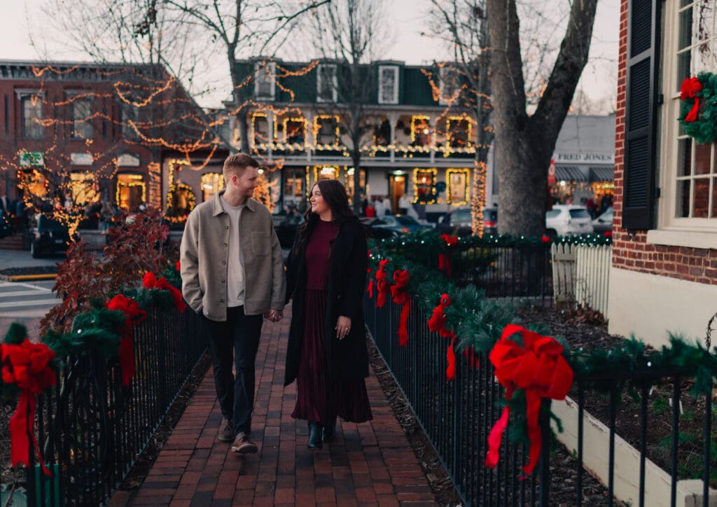 Georgia engagement photographer capturing a cozy Christmas engagement with a couple walking hand in hand through a festive town square at dusk