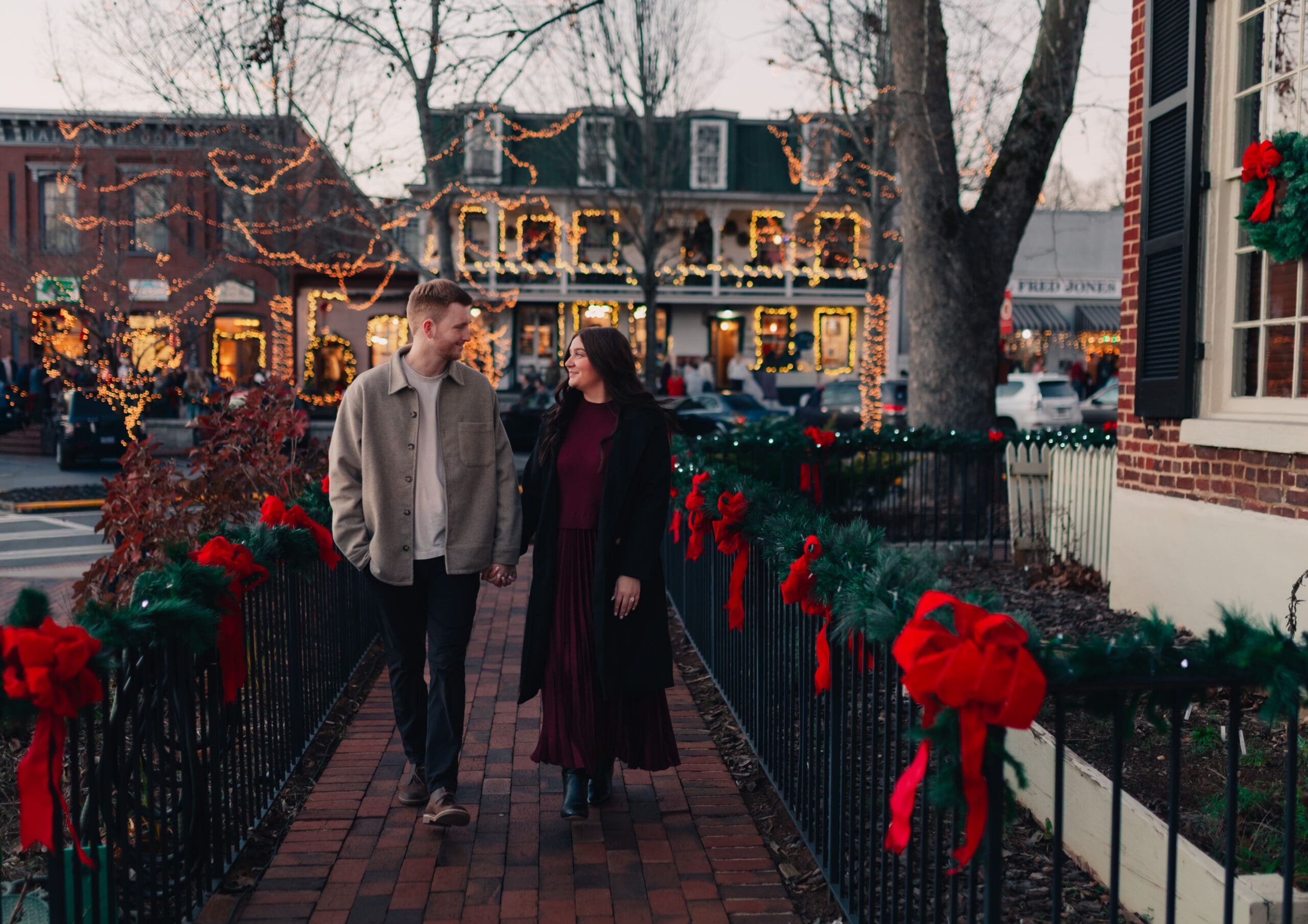 Georgia engagement photographer capturing a cozy Christmas engagement with a couple walking hand in hand through a festive town square at dusk