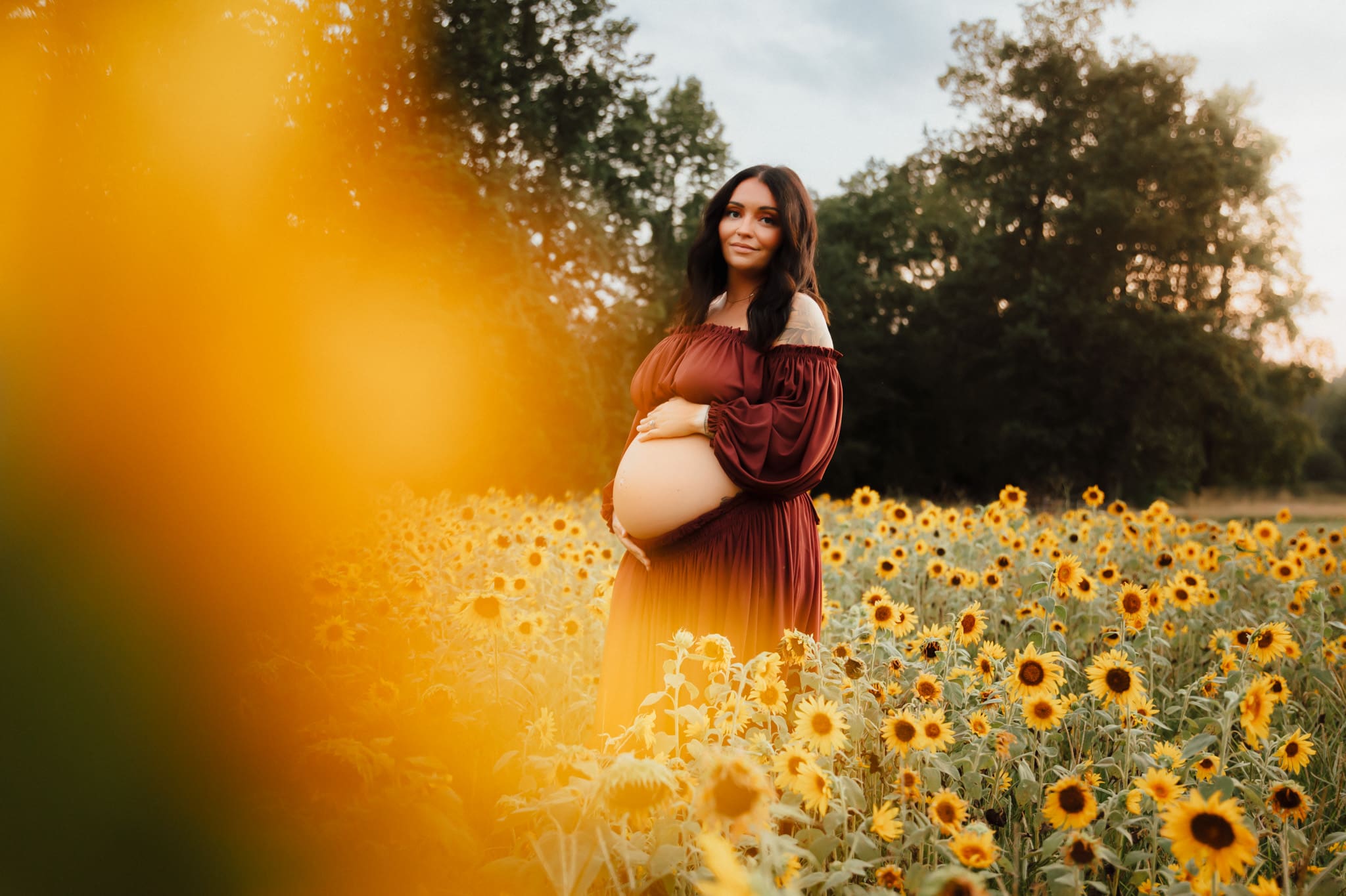 Pregnant woman standing in a sunflower field during a maternity session in Lawrenceville, Georgia, wearing a burgundy dress and gently holding her baby bump at golden hour.