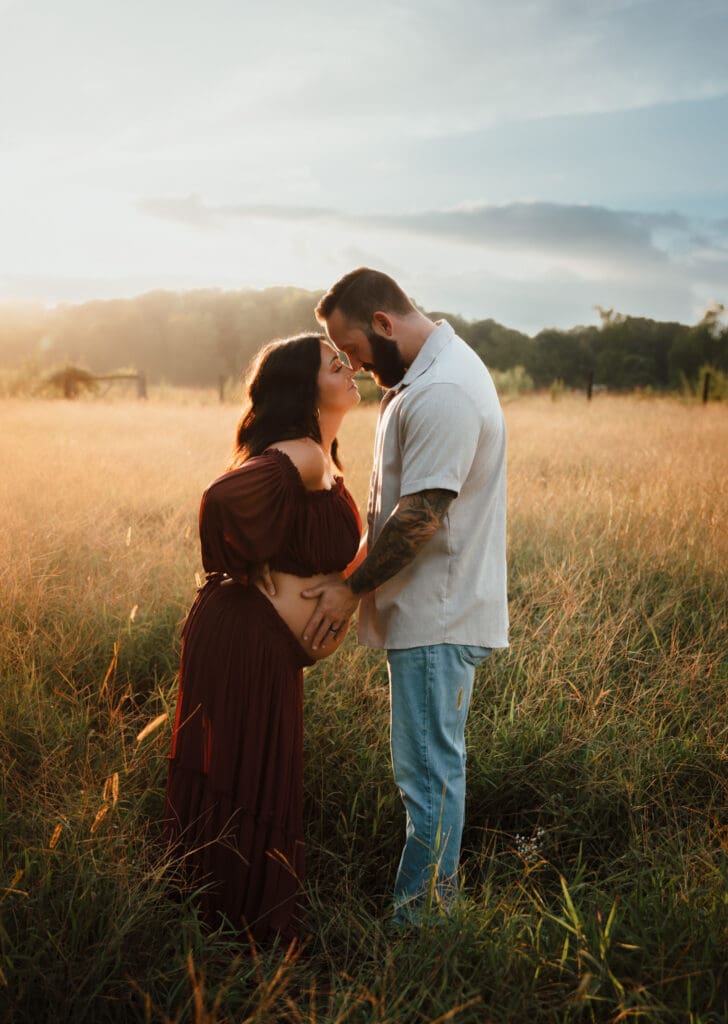 Expecting couple standing forehead to forehead in a golden field at sunset, mama wearing a deep burgundy dress and holding her baby bump.