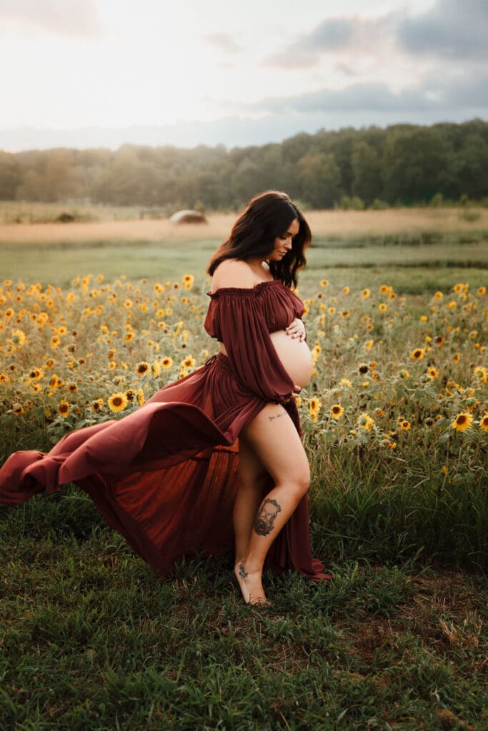 An expecting mama standing in a sunflower field, deep burgundy dress flowing in the breeze as she gently holds her baby bump.