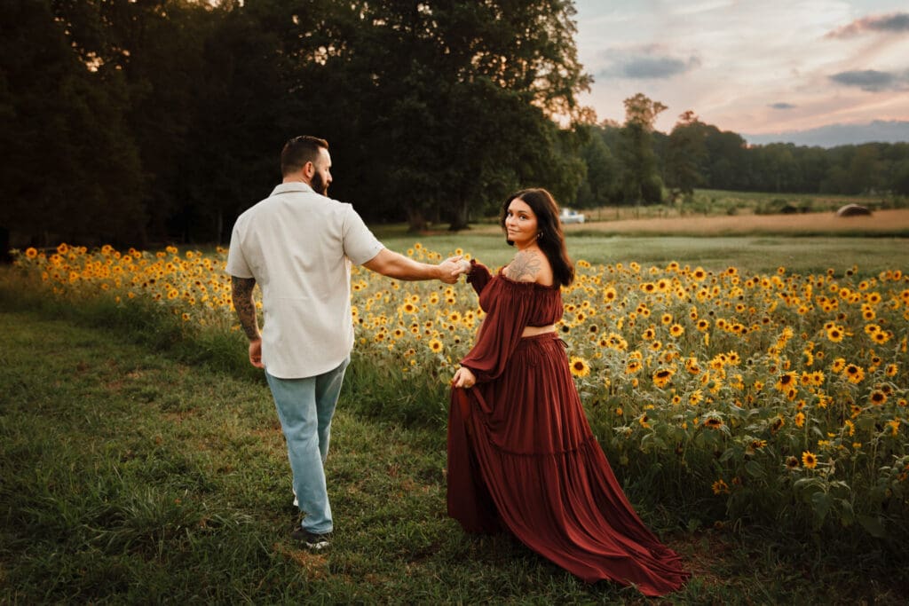 Sunflower maternity session at Prospect Farms in Lawrenceville GA with expecting parents walking hand in hand, mama in a burgundy gown surrounded by blooming sunflowers.