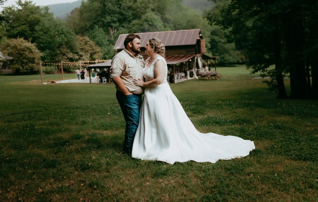 Bride and groom sharing a quiet kiss in an open grassy field with a rustic barn behind them, captured in a relaxed documentary wedding photography style in Georgia.
