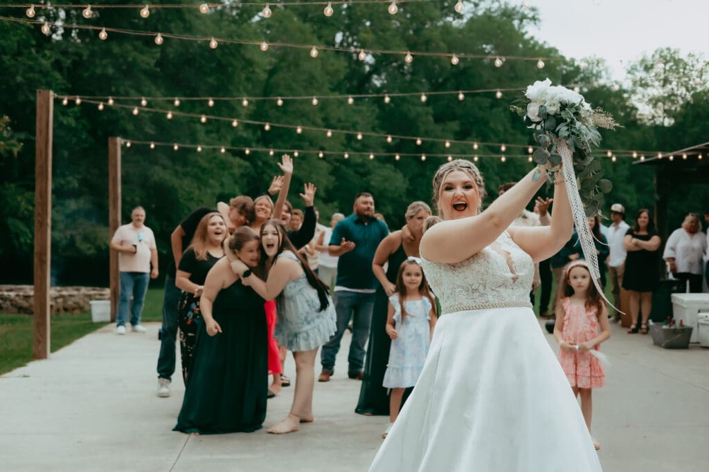 Bride tossing her bouquet during an outdoor barn reception in North Georgia, surrounded by cheering guests and string lights, captured in a candid documentary style.