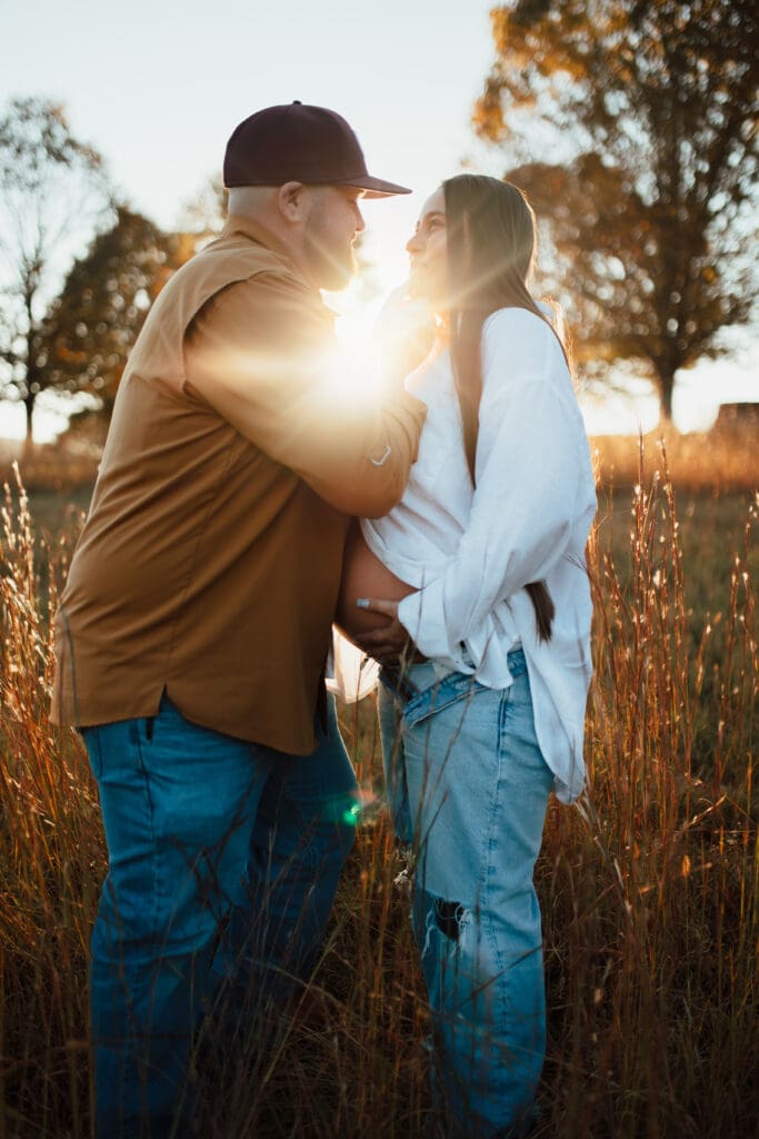 Couple standing in a golden field at sunset, sharing a quiet moment while holding baby bump