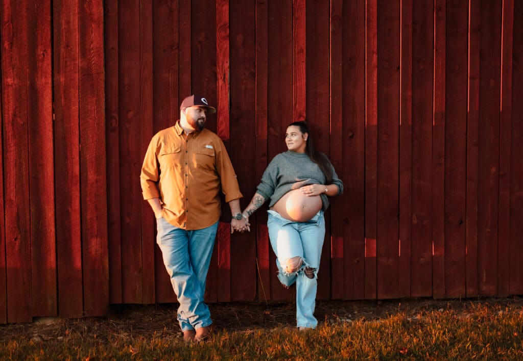Maternity couple holding hands in front of a red barn, relaxed and connected during pregnancy session