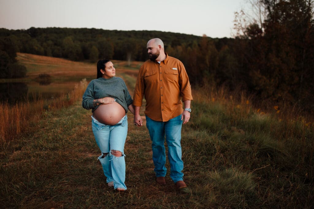 Expectant couple walking hand in hand through a grassy field, documenting their season.
