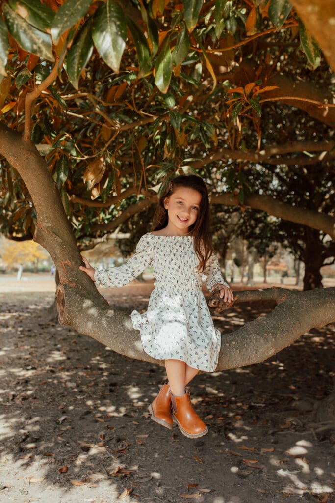 Little girl sitting on a low tree branch during a fall motherhood session, wearing a cream patterned dress and brown boots, smiling softly beneath autumn leaves.