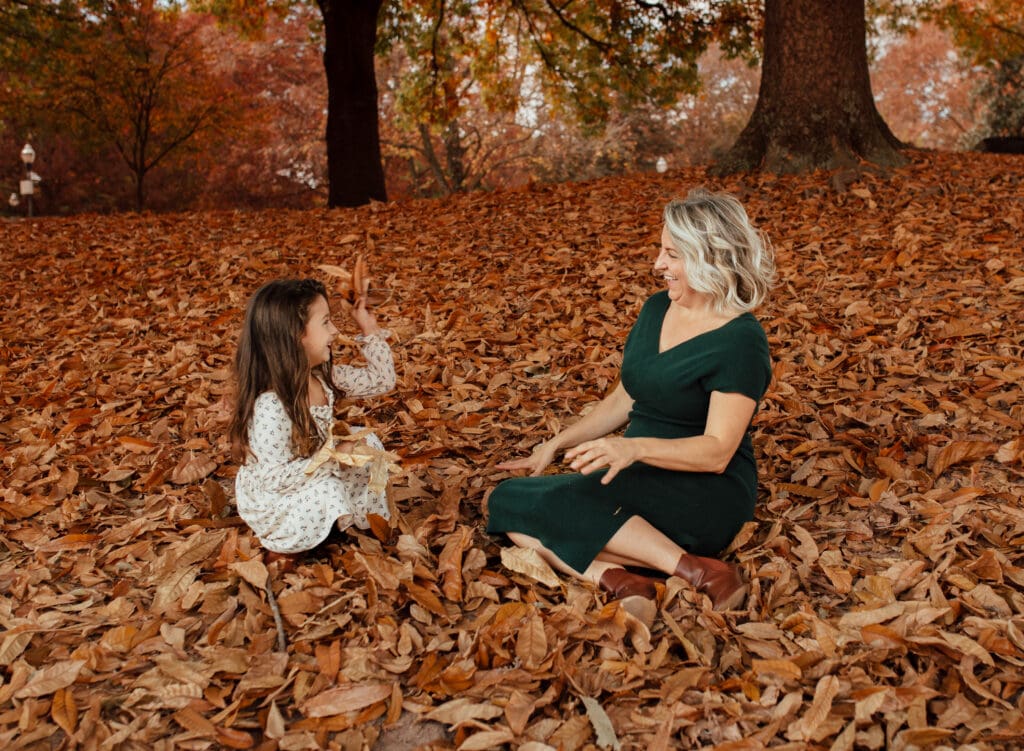 Mother and daughter sitting together in a bed of fall leaves, laughing and playing during an emotional motherhood photography session.