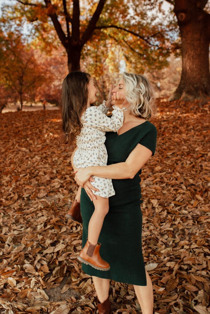 Mother lifting her daughter into an embrace as the child gently holds her face, showing tender connection surrounded by autumn color.