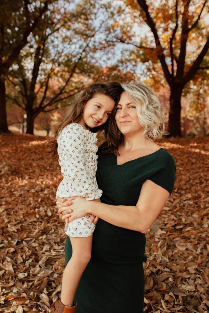 Mother holding her daughter close during a fall session, leaves covering the ground and trees glowing in warm seasonal tones.