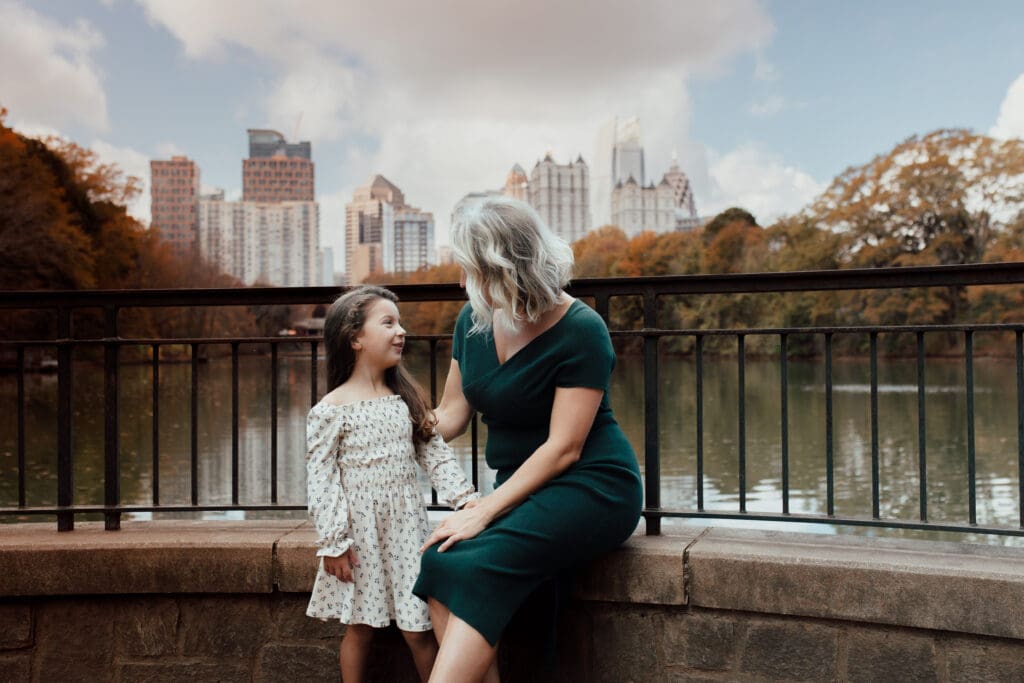 Mother sitting beside her daughter along a stone railing overlooking the lake, sharing a quiet conversation during a heartfelt motherhood session.