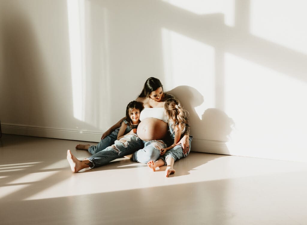 Cumming Georgia maternity photography at Wildhaven Studio featuring a pregnant mother sitting on the floor in warm natural window light with her two daughters snuggled beside her during a motherhood session.