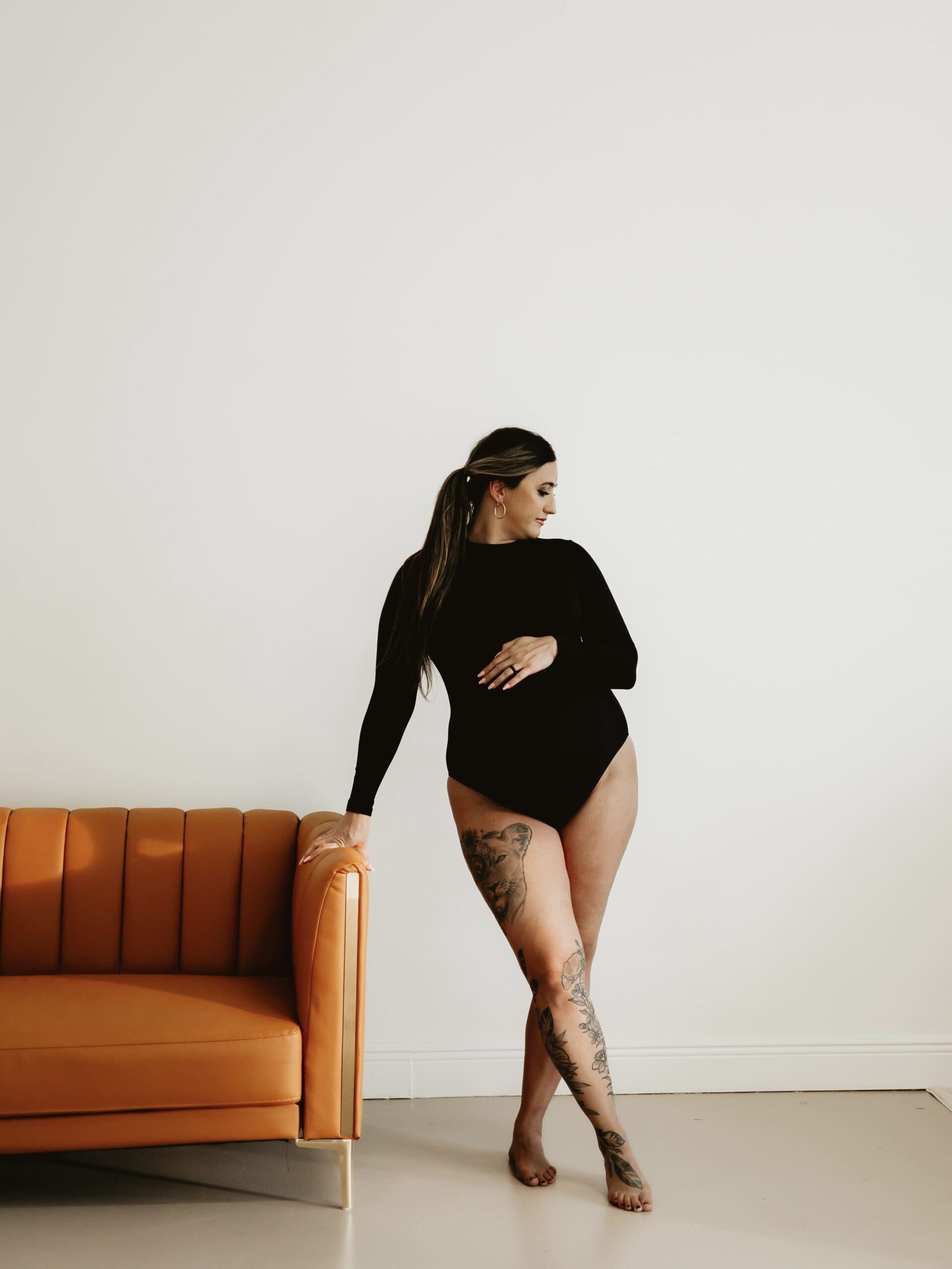 Cumming Georgia maternity photography portrait of a pregnant mother in a black bodysuit standing beside an orange chair at Wildhaven Studio.