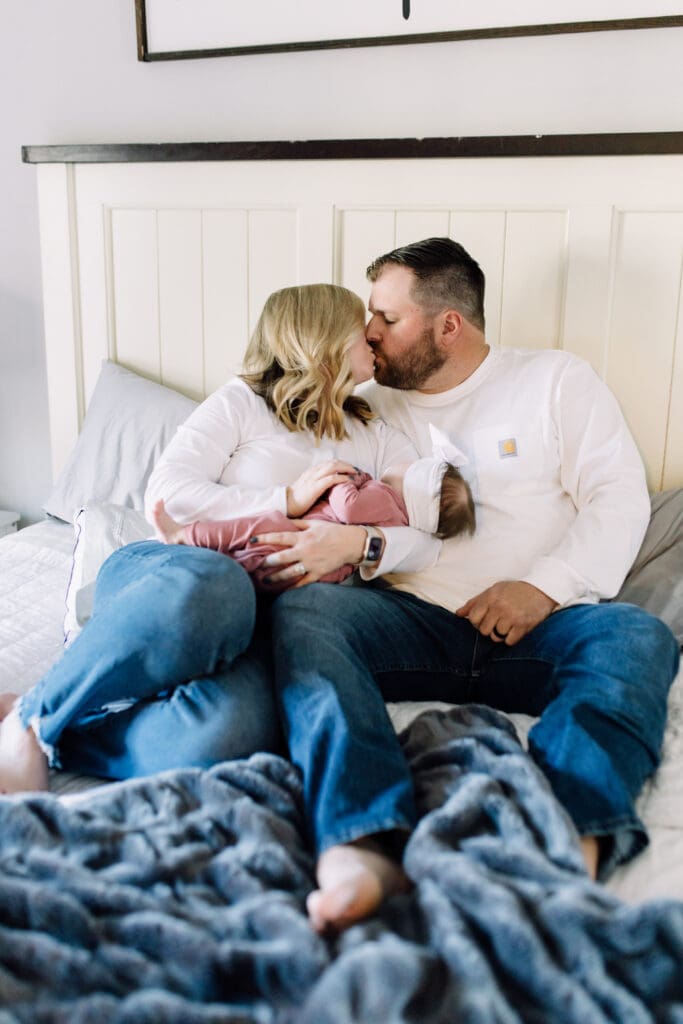 Parents kissing while holding their newborn baby girl on a bed during an in-home newborn session in Cumming, Georgia.
