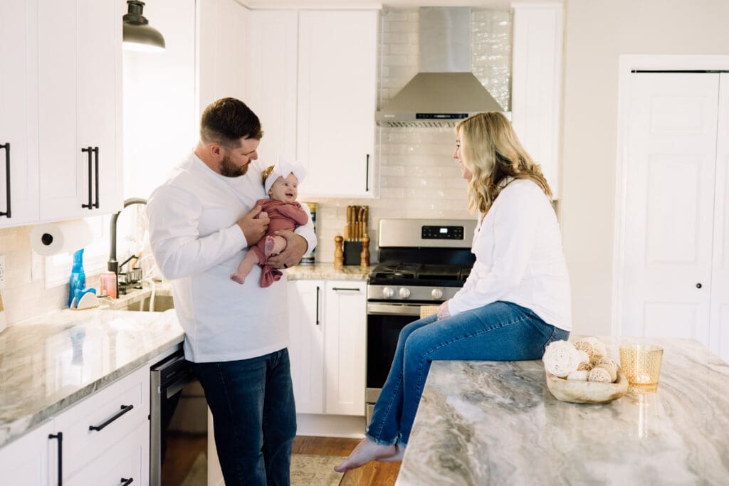 Dad holding his baby girl in a bright kitchen while mom sits on the counter during a lifestyle newborn session.