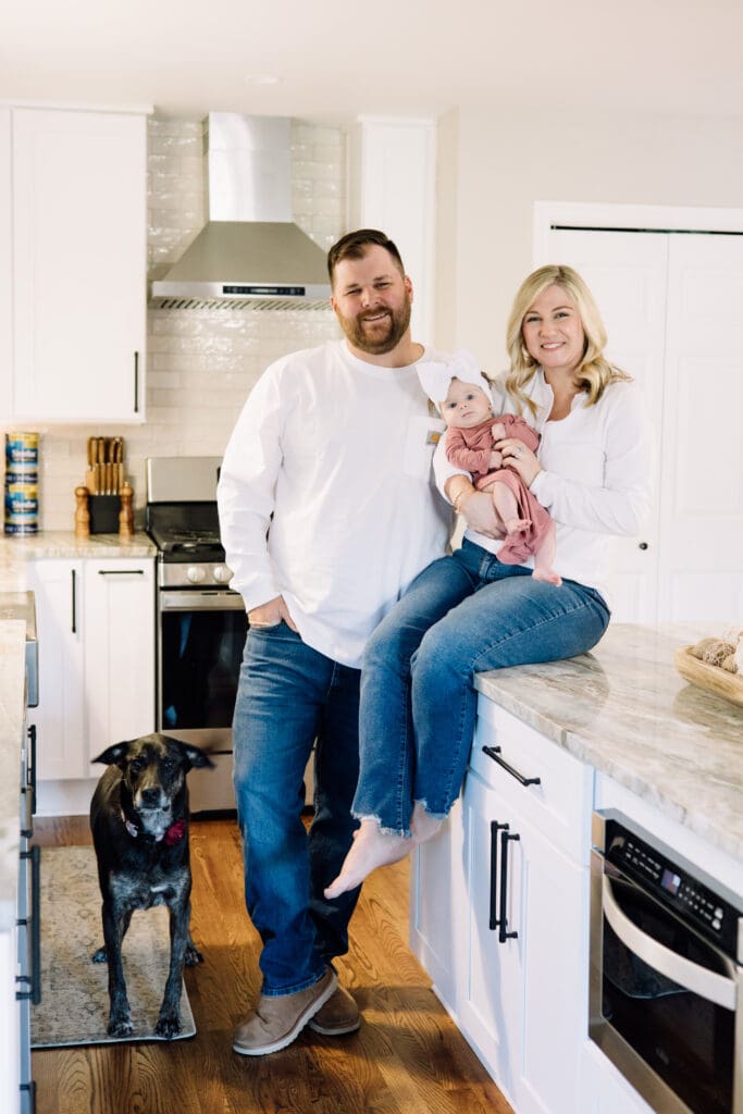 Family portrait of mom, dad, baby girl, and their dog in a bright white kitchen during an in-home newborn session. 