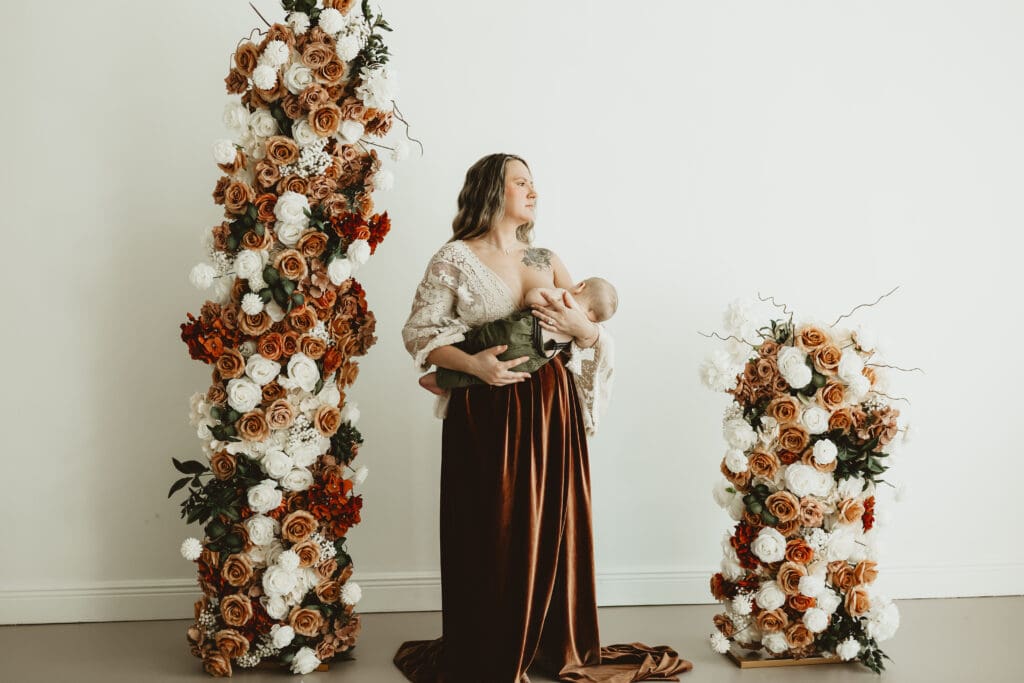 Mother holding and nursing her baby between floral pillars during a photography session at Wildhaven Studio.